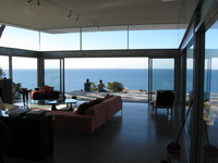 View from inside a largely glass building, furnished in a modern style.  Outside is a wooden desk with some people sitting on it, then the sea and sky.