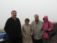 Four cold-looking people huddle on an open terrace, in the background is a white sky.