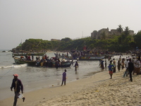 A beach crowded with people and fishing boats.  A stone fort sits on a hill above and behind the beach.