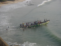 Boys pass metal basins on to a fishing boat as it moors, other boys are wading back up the beach.