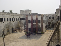 A two-storey building sits in a castle courtyard, some tourists stand in front.  Construction materials clutter the courtyard.