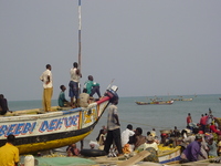 Men sit on the prow of a beached fishing boat, looking out to sea.  Below them men and women unload boats and sort fish.