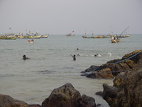 Young boys swimming in the sea.  Behind them are moored fishing boats, in the foreground are rocks.