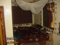 A bed built into an alcove, with shuttered windows on two sides and a mosquito net above.  The walls are decorated with ethnic motifs.