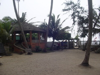 On the left, an open-sided thatched building with stairs leading up to it.  To the right is a gate in a wall.  Palm trees are scattered around the sandy foreground.