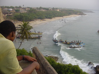 A beach curves round the top of the picture, fishing boats moored on the beach and coming in.  In the foreground a young man in a green shirt leans on a stone parapet.