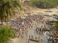 A colourful crowd on a beach, carrying basins of fish from fishing boats on the right-hand side of the picture.