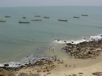 Small boys with makeshift surfboards (planks) swimming in a bay, behind them fishing boats are moored.  In the foreground a rocky and sandy beach.