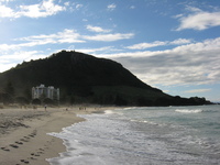 A hill (silhouetted against the sky) rises behind some tall apartment buildings, below is a sandy beach.