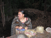 A Maori woman in a cloak peels sweet potatoes using a mussel shell.
