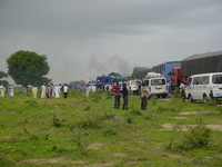 A queue of vehicles parked all over the road, people standing around. Black smoke rising in the background.