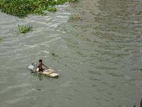 A muddy river with lumps of vegetation floating downstream.  On a dodgy-looking raft sits a small boy, paddling with flip-flops on his hands.