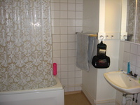 A white-tiled bathroom.  A plastic shower curtain screens the bath, to the right is a sink with a mirrored cabinet over it.