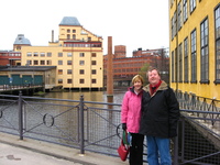 My parents standing on a bridge in front of part of a river, surrounded by former industrial buildings.  Mum and Dad are wearing big warm coats.