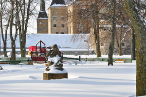 In the background part of a castle is lit by winter sun.  In front of it a childrens' playground sits among trees and snow.  In the foreground a statue of a woman looks down at the snow, her face lit by the reflected light.