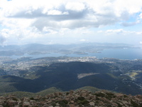 Looking down from a mountain onto a city built along the side of a bay.  A bridge crosses at a narrow point and hills rise on the far side.