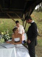Hannah sits on a bench in a wooden shelter to sign the wedding register, Lee is standing behind her.