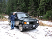 Dugly stands in front of a mud spattered Land Rover Discovery.  Snow lies on the ground and there are trees all around.