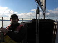 Adrian to the left of picture, in the cockpit of a yacht, against the sky.