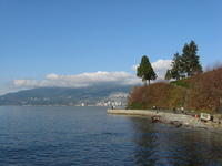 A path with pedestrians leads from the bottom right to the centre of the picture, above it and to the right is a bluff with a few trees.  In the background hills covered in trees and houses rise from the water.