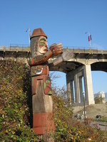 A carved wooden totem pole with outstretched arms stands in front of a high concrete bridge.