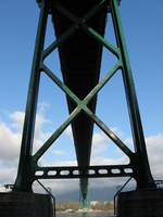 Under Lions' Gate bridge. A green-painted suspension bridge is directly overhead going from top to bottom of the picture. The bridge is seen between the spars of one of its towers.