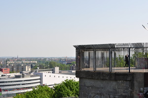 An ugly concrete structure forms a large octagonal platform surrounded by an uglier metal fence.  A man stands on the platform, apparently doing some kind of exercise.  To the left, past the platform, a modern shopping centre can be seen.