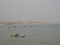 A cityscape on in the middle distance, with tall buildings and cranes.  In the foreground are colourful fishing boats on the sea.