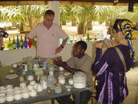 A Ghanaian man sits at a table covered in ceramic molds and jars of powdered glass, explaining bead making to people standing around the table.