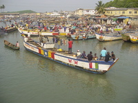 Fishermen stand and sit in a brightly-coloured boat passing other, moored, boats on its way into the harbour.  'In Him is life' is painted on the side.