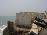 Waves crash on rocks below a whitewashed castle wall.  On the right: rusty cannons point out to sea.