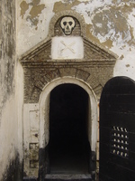 The entrance to a dark cell with a thick wooden door.  A skull and cross bones are carved above the doorway.