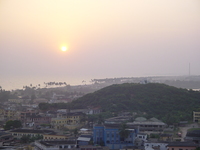 The sunsets above a tree-covered hill with a small whitewashed fort on top.  Below the fort is a jumble of painted buildings.