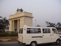 A blocky triumphal arch with large black star on top and a packed minibus hurtling past in front.