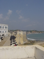 In the foreground: castle battlements and cannons.  Behind: buildings on a hill rising up from the sea.  To the left: whitewashed castle buildings.