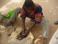 A man sits on the ground, polishing glass beads in a hollowed-out stone, using water and sand.