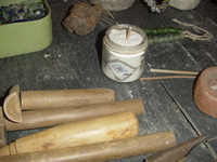 A glass jar containing coloured powder arranged in a diamond-shaped pattern sits on a table among wooden tools, glass and beads.