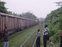A few people standing around the tracks beside a train.  Another train sits further up the tracks.