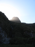 Dark mountains and sky, the sun is shining in front of one peak, lighting up the haze.