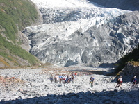 A line of tourists walking across rocks in front of a glacier face.