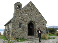 A small stone church in front of a lake and mountains.