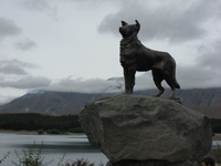 A statue of a sheepdog in front of a lake and mountains.