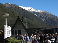 A train stopped in a small station below snow-capped peaks, passengers fill the platform.