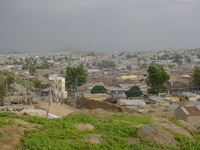 In the foreground a hill slopes down towards the remnants of a mud wall and a modern white concrete gate with a major road passing through it.