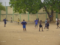 Young men playing football on a dirt pitch in front of a high wall.