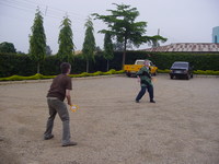 Two men playing tennis with children's bats on a gravel car park
