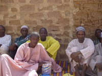 A group of men sitting on mats in front of a mud-brick wall.