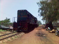 A scruffy-looking locomotive on a single-track line passing trees and dusty waste ground.