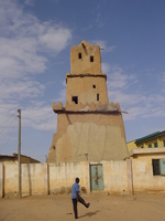 A three-storey mud building behind a concrete wall.  A boy is playing football in front.