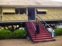 A wooden colonial house on stilts.  A man stands on the red painted steps.